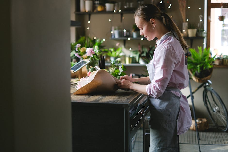 A young female florist, dressed in a pink shirt and grey apron, is carefully arranging a bouquet of fresh flowers on a wooden counter inside a cozy flower shop. The bouquet features a mix of pink rose