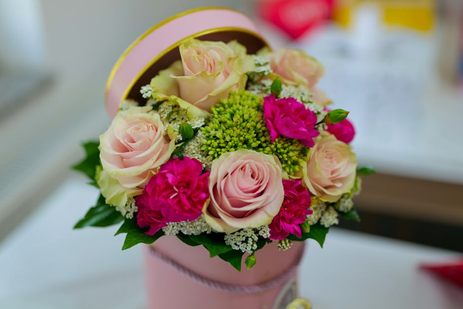 A floral arrangement featuring soft pink roses with fully bloomed petals, complemented by vibrant fuchsia carnations, and clusters of small white and green filler flowers, all arranged in a round pink